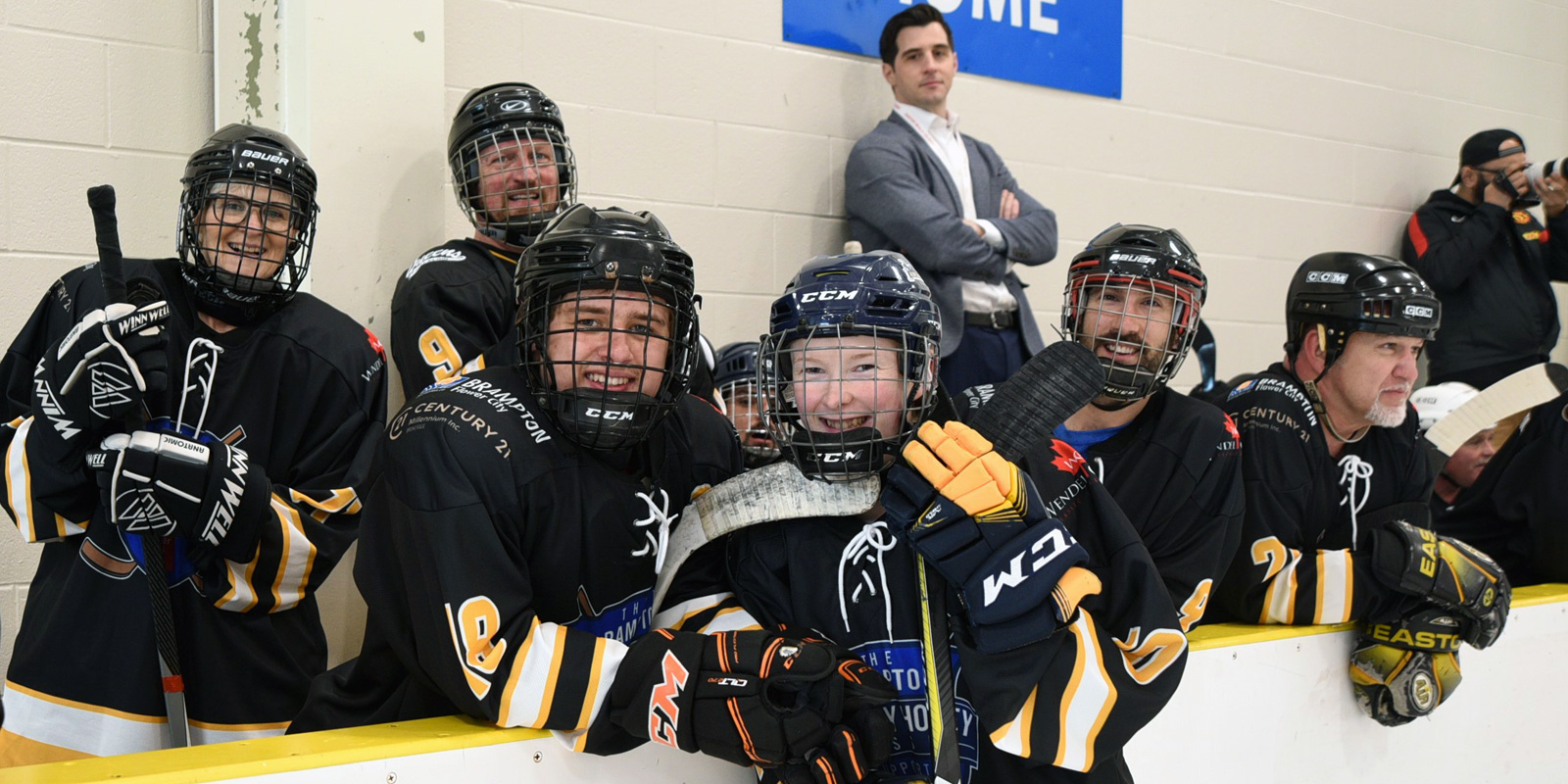 A team in balck jerseys pose in front of the bench with a coach in behind leaning against he brick wall